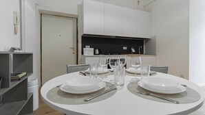 A dining area with a round table set for two, white plates, clear glasses, and cutlery. There is a bookshelf to the left and a white door in the background. - Italianway - Sigieri 10 (Milan)