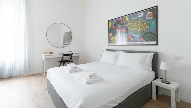 A hotel room with a bed, a desk, a chair, a round mirror, a bedside table with a lamp, and a large framed picture on the wall. - Italianway - Sigieri 10 (Milan)