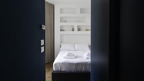A modern hotel room with a bed, white linens, a built-in shelf unit, and a wooden floor.
