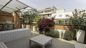 An outdoor patio with a tiled floor, a white table, and chairs. There are potted plants and a pergola providing shade.