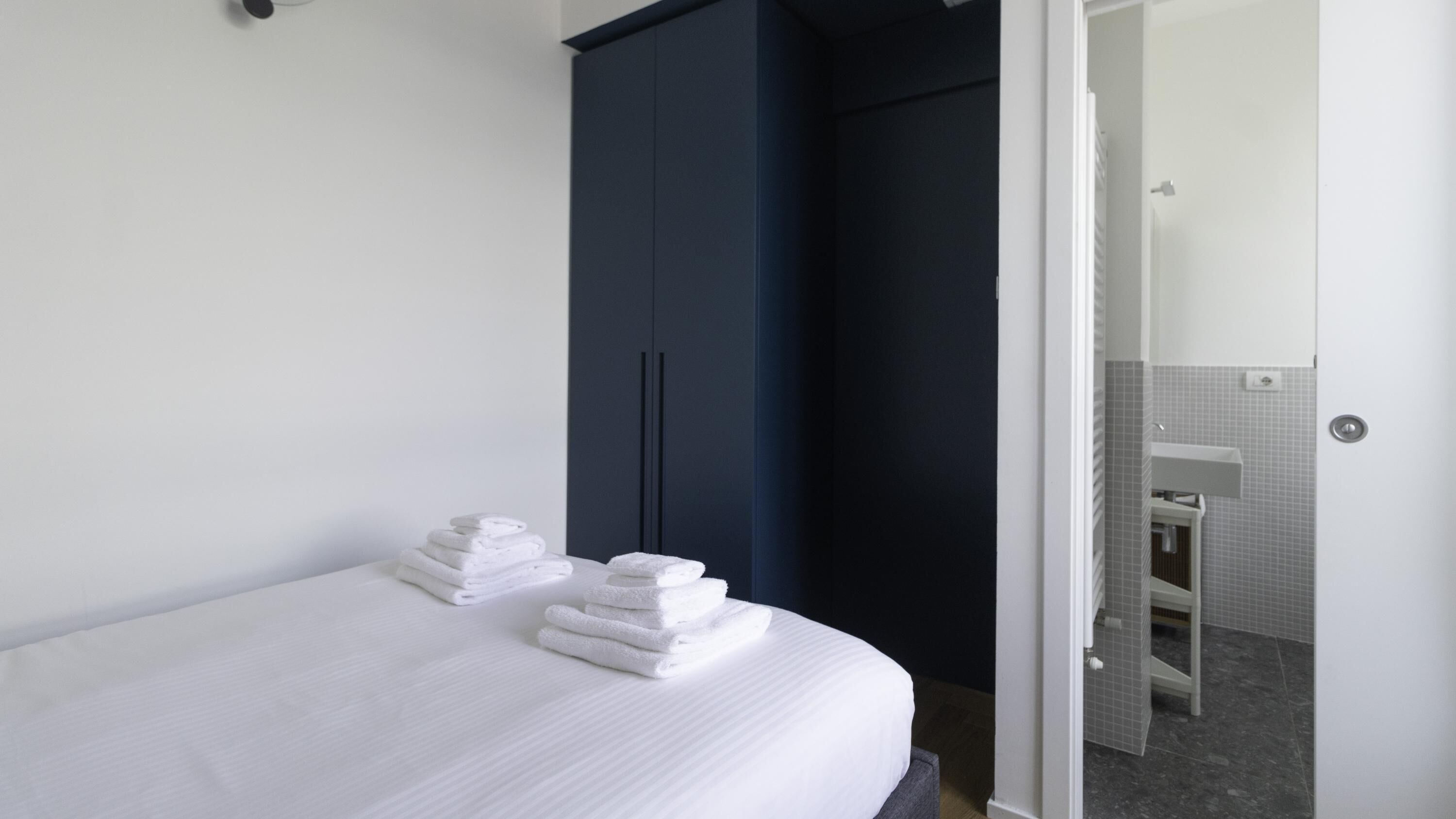 A hotel room with a neatly made bed, a dark blue wardrobe, and a bathroom visible through an open door.