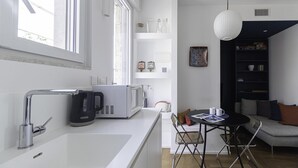 A modern kitchen with a white countertop, a coffee maker, a microwave, and a kettle. There is a dining area with a table and chairs, and a bookshelf in the background.