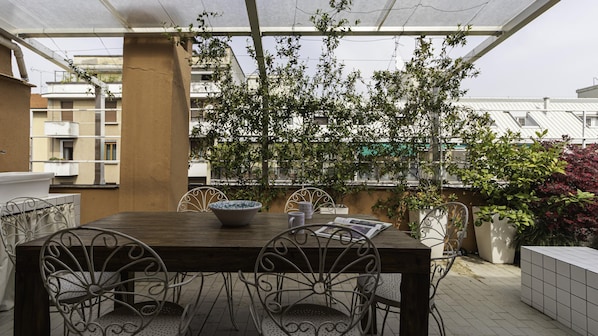 An outdoor patio with a wooden table and metal chairs, surrounded by potted plants and a view of buildings.
