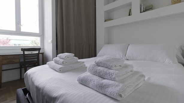 A neatly made bed with white linens and towels, a wooden desk, a chair, and a window with curtains.