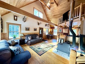 A cozy cabin interior with a living area, a fireplace, wooden beams, and a loft bedroom.