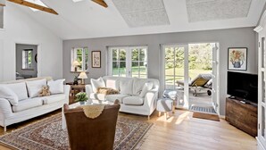 Amplia sala de estar con un sofá blanco, un sillón marrón y un televisor. Hay ventanas grandes y una puerta que da a un área exterior.