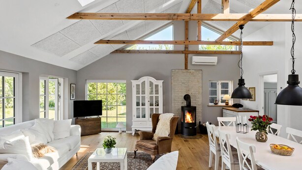 Un loft espacioso con techos altos, vigas de madera a la vista, chimenea, comedor y ventanas amplias con vistas al verdor.