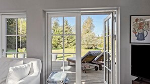 Una habitación con un sofá blanco, una mesita con una lámpara y un ventanal con vistas a un jardín y mobiliario exterior.