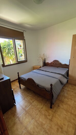 A bedroom with a wooden bed, a nightstand, a dresser, and a window with a view of greenery.