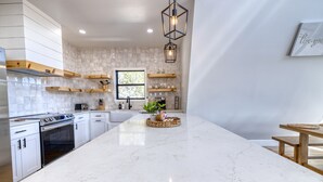 Uma cozinha moderna com armários brancos, uma ilha grande de mármore, prateleiras de madeira e um lustre.