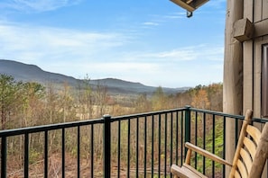 A wooden cabin with a balcony overlooking a forested valley and distant mountains.
