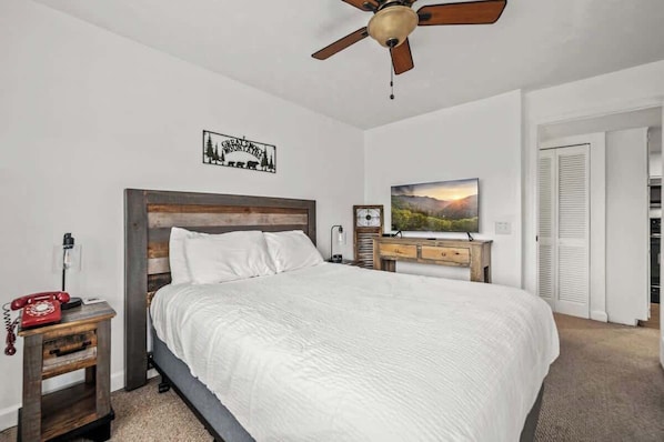 A bedroom with a bed, a nightstand with a red telephone, a wooden headboard, a ceiling fan, a wall-mounted clock, a framed picture, and a closet with white shutters.