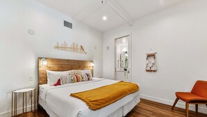 Quarto com cabeceira de madeira, roupa de cama branca, manta cor mostarda, mesa de cabeceira pequena e uma cadeira.