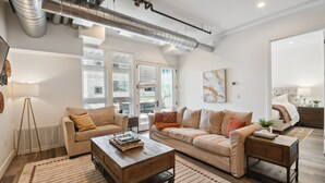 A spacious living room with a beige sofa, a wooden coffee table, and a large window with blinds. - Brushed & Bold | RiNo Art Lofts (Denver)