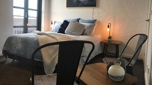 A bedroom with a bed, a chair, a table, a lamp, and a painting on the wall. - Funky Fitzroy 2 bedroom Apartment in Old Chocolate Factory (Fitzroy)