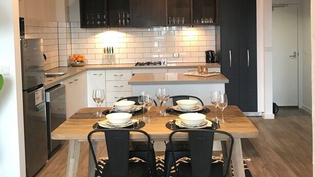 A modern kitchen with a dining area, featuring a wooden table set for four, black chairs, and a patterned rug. - Funky Fitzroy 2 bedroom Apartment in Old Chocolate Factory (Fitzroy)