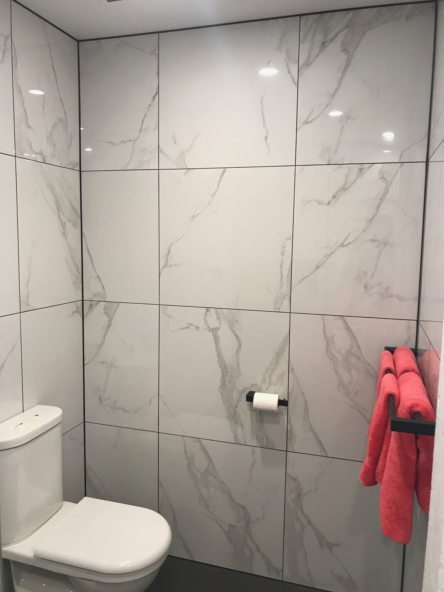 Une salle de bain carrelée de marbre blanc, avec des toilettes blanches et un porte-serviettes orné de serviettes rouges.