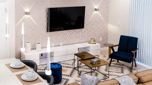 Un salon moderne avec un téléviseur à  écran plat, une console blanche, un fauteuil bleu foncé et un tapis à motifs.