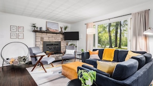 A modern living room with a stone fireplace, comfortable seating, and a large window offering a view of trees.