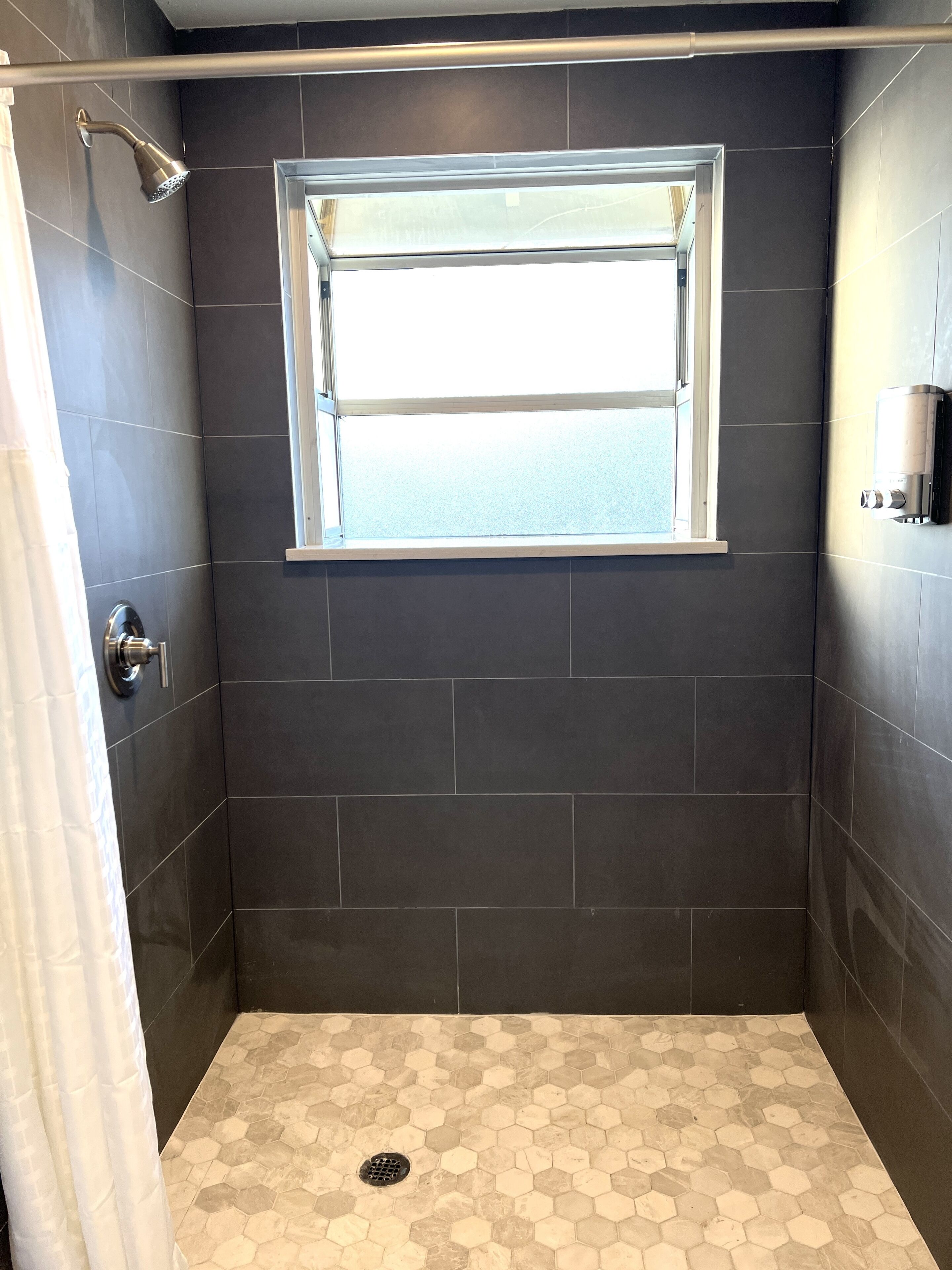 Una ducha con ventana, azulejos oscuros y suelo de mosaicos hexagonales.