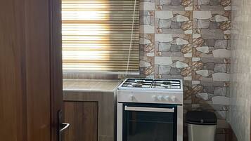 A compact kitchen with a gas stove, a trash bin, and a window with blinds.