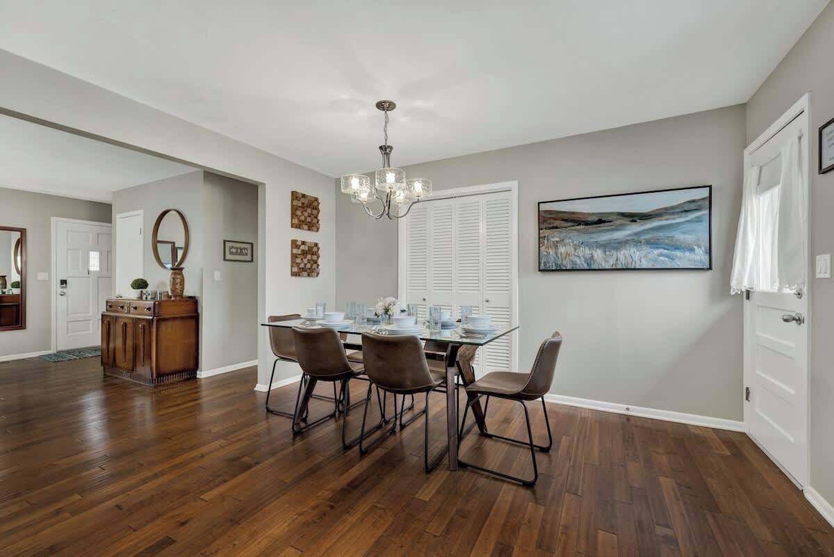 A dining area with a glass table, four chairs, a chandelier, and a painting on the wall.