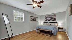 Un dormitorio con cama, ventilador de techo, lámpara de pie y ventana grande.