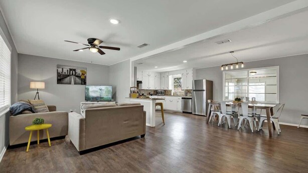 Un salón espacioso con un sofá, una mesita amarilla y un televisor. Se observa la zona de la cocina con armarios blancos y una mesa de comedor.