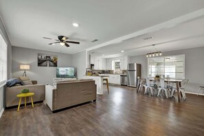 A spacious living room with a sofa, a small yellow table, and a TV. The kitchen area is visible with white cabinets and a dining table. - Grape Getaway Haus (Fredericksburg)