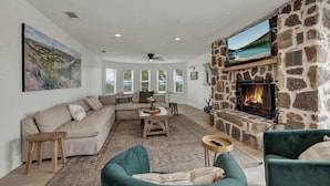 A living room with a stone fireplace, a large TV mounted above it, a sofa, and a green armchair. - Grasslands Pool Hot Tub Hill Country Views (Johnson City)