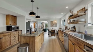 A modern kitchen with wooden cabinets, a central island, pendant lighting, and a dining area with a view. - Grasslands Pool Hot Tub Hill Country Views (Johnson City)