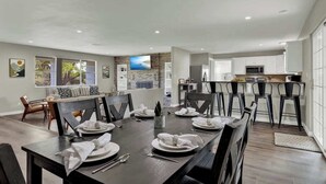 A spacious dining area with a long table set for six, surrounded by modern chairs. A kitchen is visible in the background with a bar area. - River Oaks Retreat With Hot Tub (Fredericksburg)