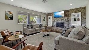 A modern living room with a stone fireplace, a grey sofa, a coffee table, and a dining area with a chair and a small table. - River Oaks Retreat With Hot Tub (Fredericksburg)