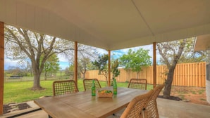 A covered patio with a wooden table and wicker chairs, overlooking a grassy area with trees and a wooden fence. - River Oaks Retreat With Hot Tub (Fredericksburg)