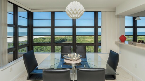 Un'elegante zona pranzo con tavolo in vetro, sedie nere e un centrotavola decorativo. Ampie finestre offrono vista sull'oceano e sul verde.
