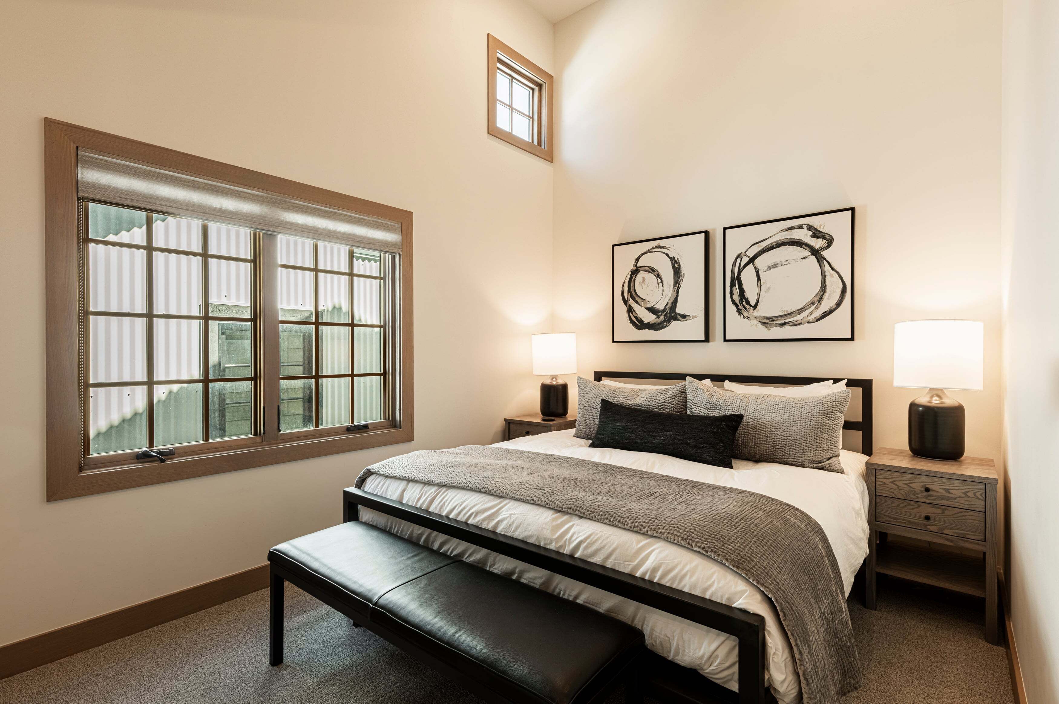 A bedroom with a large window, a bed with a bench, two bedside tables with lamps, and two abstract paintings on the wall.