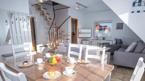 Un coin repas avec une table en bois, des chaises blanches et un saladier rempli de fruits.