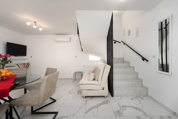 A modern living room with a glass dining table, a sofa, and a staircase.
