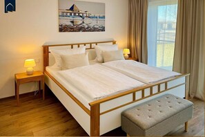 Una cama doble con estructura de madera, dos mesitas de noche con lámparas, un banco y un cuadro de un muelle en la pared.