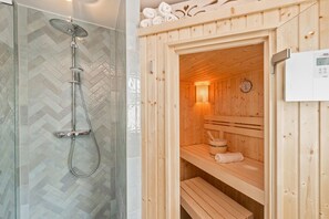 Una sauna con bancos de madera y un calentador fijado a la pared, junto a una zona de duchas con puerta de vidrio.