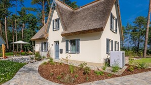 Une maison à deux étages, avec un toit de chaume, une allée pavée et un jardin où poussent différentes espèces de plantes.