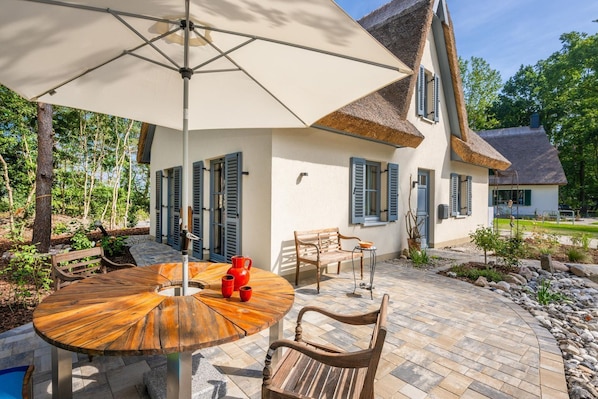 A patio with a round wooden table, two wooden chairs, a red pitcher, and a white umbrella.