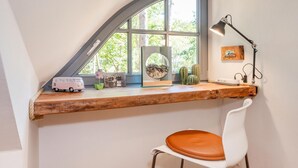 Un espace de travail comprenant un bureau en bois, une chaise blanche, une lampe et une fenêtre donnant sur des arbres.