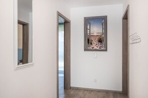 Een gang met een houten vloer, een deur die naar een andere kamer leidt, en een aan de muur hangend schilderij van een strandgezicht.