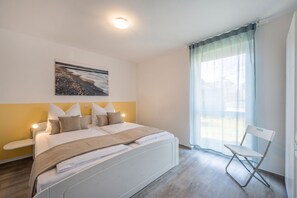 Een slaapkamer met een bed, een stoel, een nachtkastje, een raam met gordijnen en een aan de muur hangend schilderij.