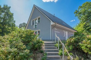 A two-story house with a grey exterior and a dark roof, surrounded by lush greenery and a staircase leading to the entrance.