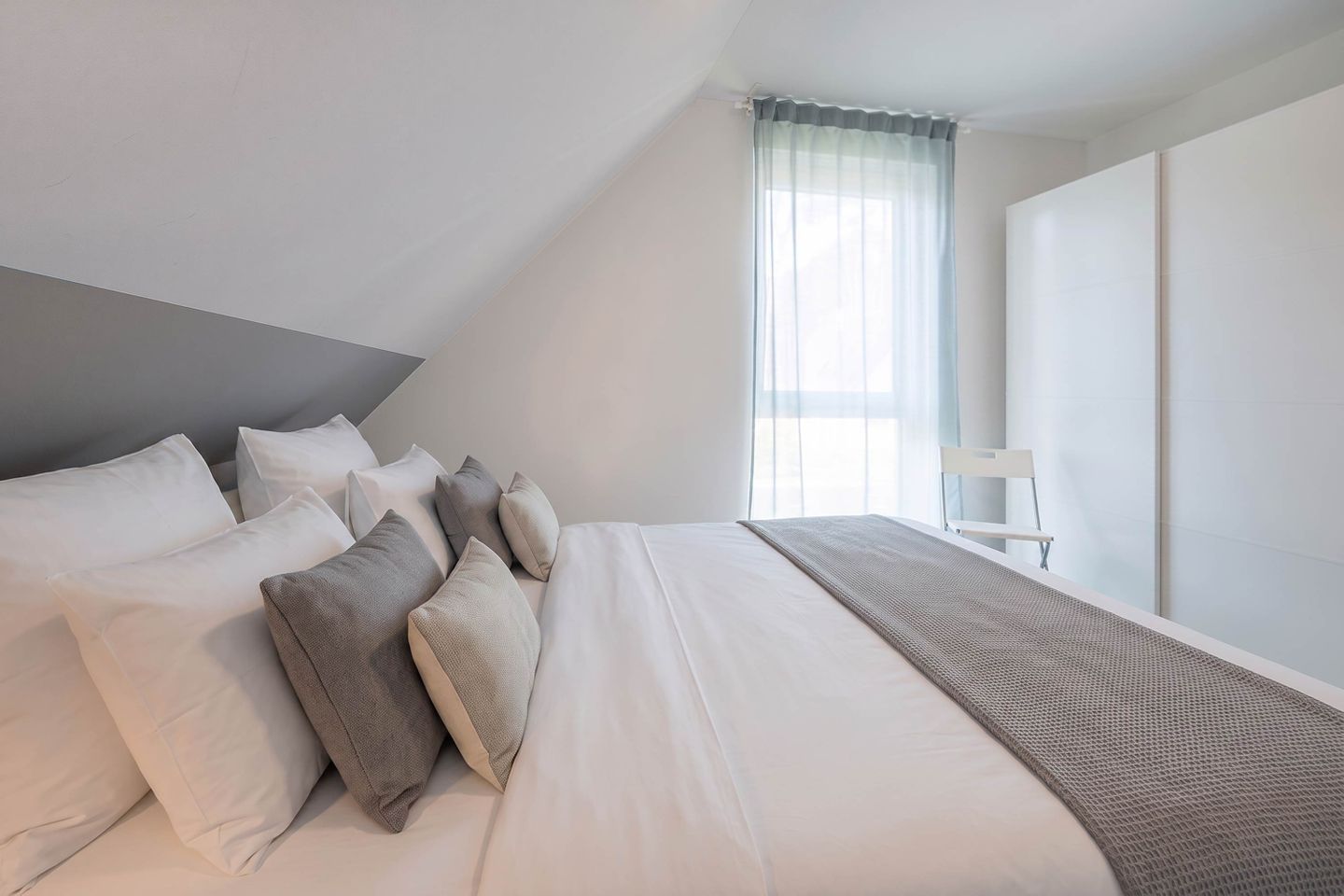 Une chambre moderne avec un grand lit, de nombreux oreillers, un fauteuil et une fenêtre ornée de rideaux légers.