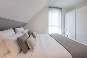 A modern bedroom with a large bed, multiple pillows, a chair, and a window with sheer curtains.