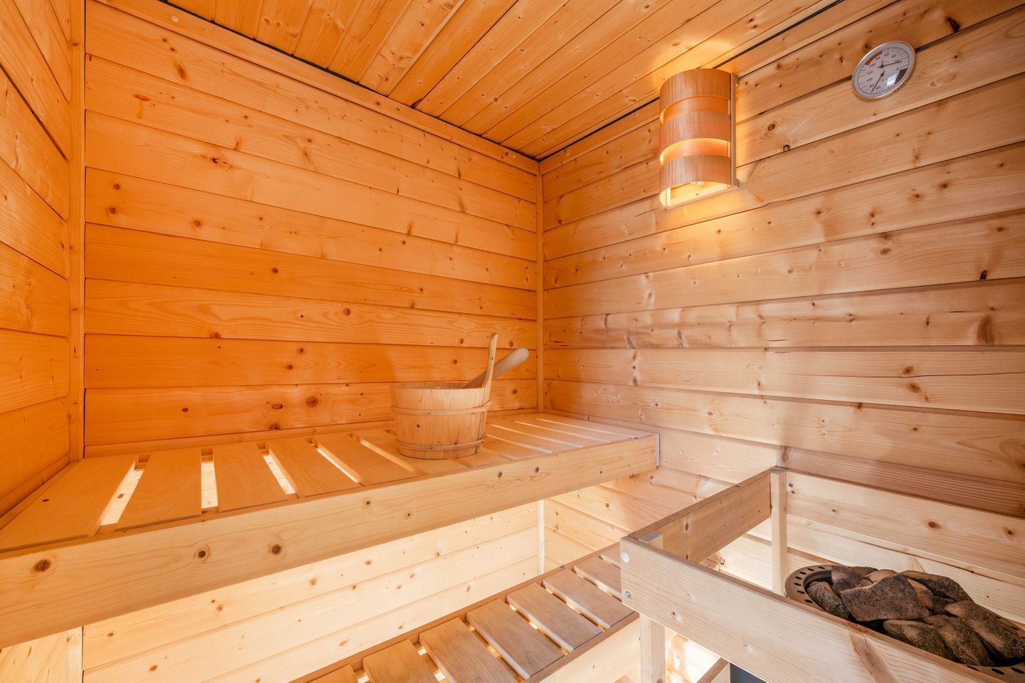 A wooden sauna with benches, a bucket, and a wall-mounted thermometer.