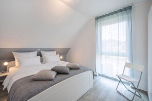 Une chambre moderne avec un lit, des lampes de chevet, un fauteuil et une grande fenêtre ornée de rideaux légers.